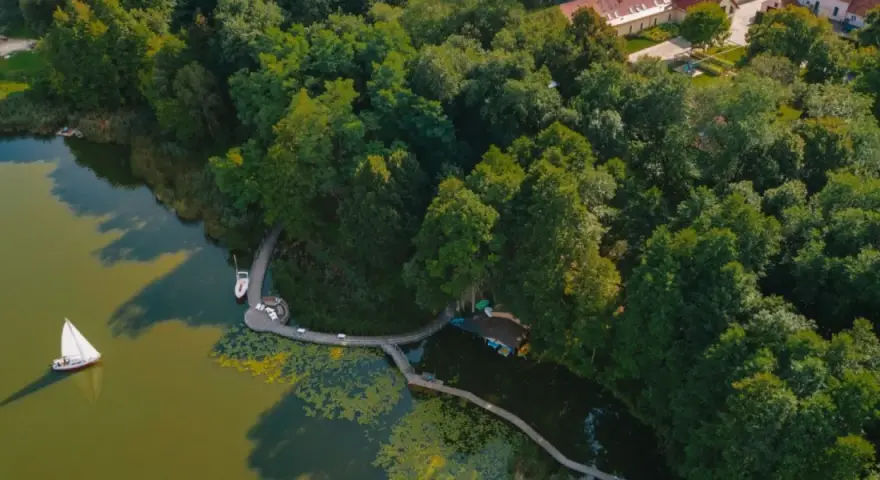 Żaglówka i deska SUP na spokojnym jeziorze ostrowskim, otoczonym bujną zielenią i drewnianą kładką.
