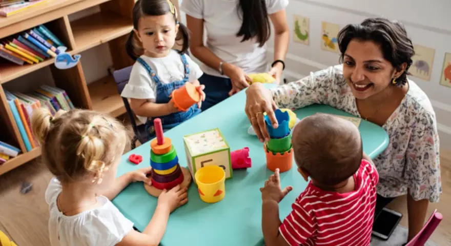 Zwei Frauen und drei Kleinkinder spielen mit bunten Spielsachen. Eine Frau hilft einem Kind, Türme zu stapeln, was eine tolle Methode ist, um Kinder zweisprachig zu erziehen.