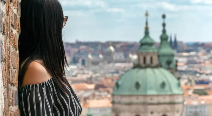 Vista di una donna con capelli scuri appoggiata a un muro di mattoni, con lo sguardo rivolto verso un panorama urbano di **città europee da visitare in 3 giorni**.