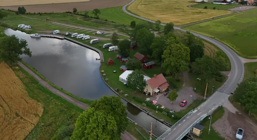 Jonsboda camping vid en kanal med husbilar, båtar och röda stugor. En bro leder över vattnet.