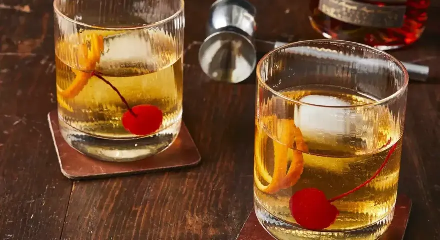 Klasyczny Old Fashioned przepis: dwa drinki z lodem, skórką pomarańczy i wisienką, obok miarki barmańskiej i butelki bourbona.