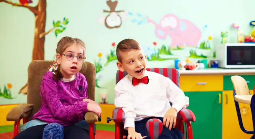 Dwoje dzieci w fotelikach, dziewczynka w okularach i chłopiec w muszce, na tle kolorowej ściany z rysunkami.