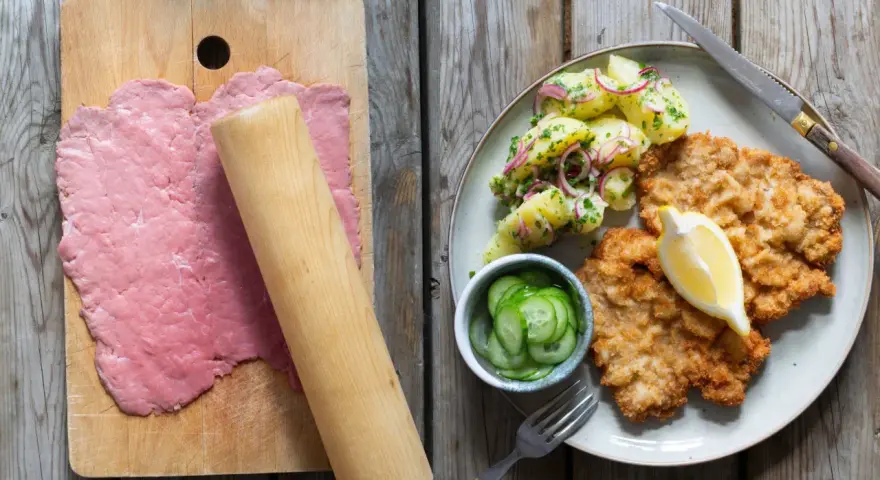 Förberedelser för schnitzel recept: tunt bankat kött på skärbräda, bredvid en tallrik med schnitzel, potatissallad och gurka.