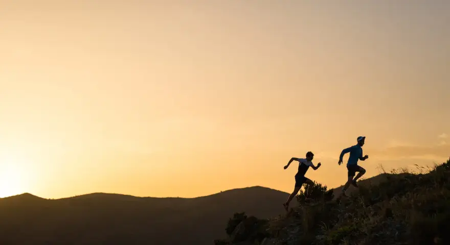 Dos corredores se enfrentan a una exigente carrera de senderos al amanecer, sus siluetas recortadas contra un cielo anaranjado.