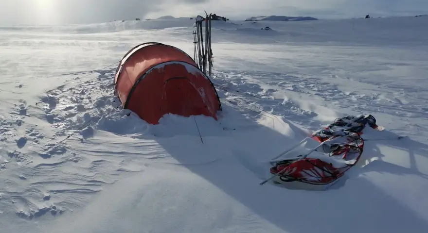 Ein rotes Wintercamping Zelt steht im Schnee, daneben Skier und ein Schlitten. Die Landschaft ist weit und verschneit.