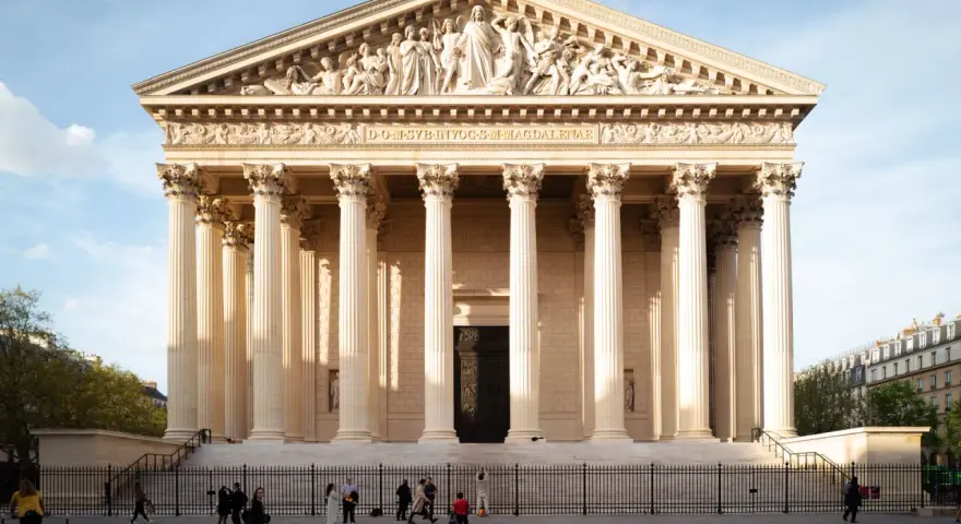 La imponente iglesia de la Madeleine en París, con sus columnas neoclásicas y esculturas en el frontón, se alza bajo un cielo azul.