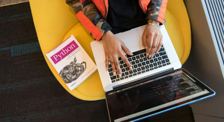 Osoba w pomarańczowej kurtce pisze na laptopie obok książki o Pythonie, siedząc na żółtym krześle.