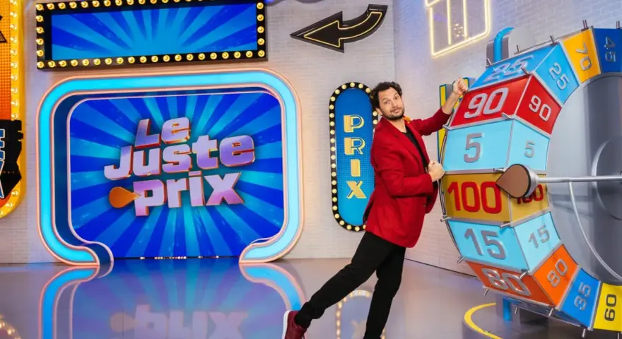 Un animateur pose près de la roue du jeu "Le Juste Prix". L'illustration du plateau est colorée et lumineuse, promettant de beaux gains.