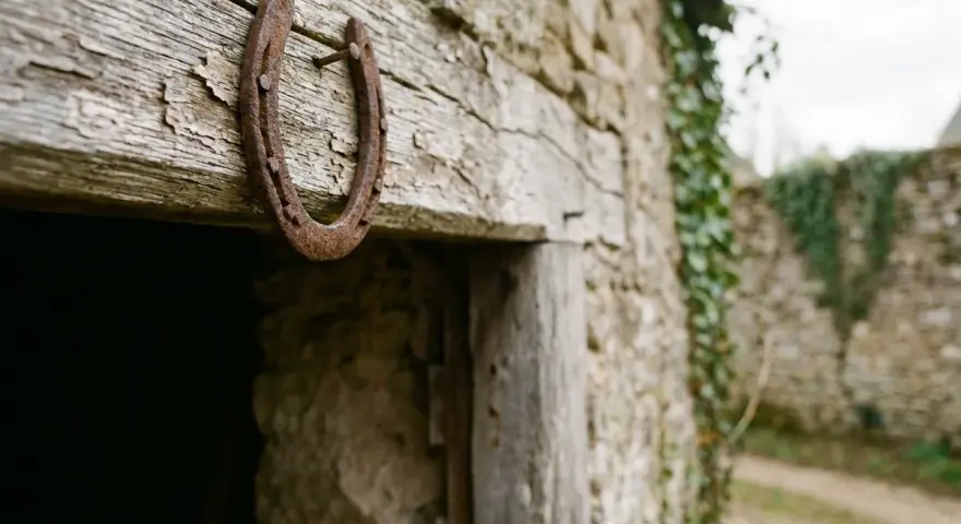Un vieux fer à cheval pointes en bas, rouillé, est cloué sur une poutre en bois patiné, devant une bâtisse en pierre recouverte de lierre.