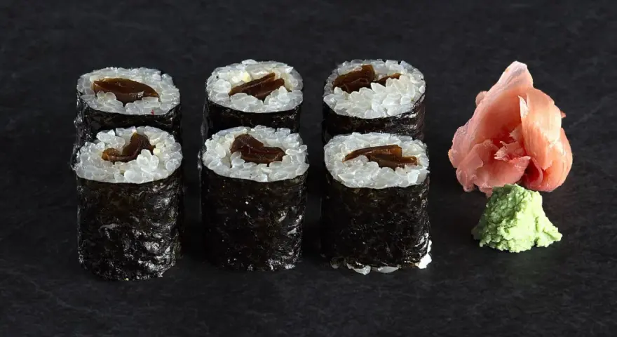 Sześć roladek sushi z ryżem i ciemnym nadzieniem, prawdopodobnie sushi z tykwą. Obok różowy imbir i zielone wasabi.