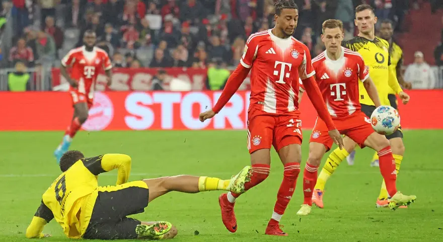 Intensywna akcja na boisku podczas meczu składy: Borussia Dortmund – Bayern Monachium. Piłkarz Bayernu w czerwonej koszulce próbuje odebrać piłkę leżącemu zawodnikowi BVB.