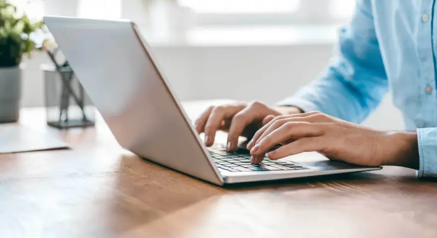 Person tippt auf einem silbernen Laptop auf einem Holztisch.