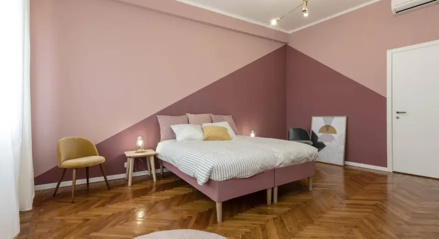 Camera da letto con pareti bicolore, un letto rosa, una sedia gialla e un parquet a spina di pesce. Un esempio di come pitturare una stanza con stile.