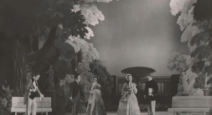 Scena z przedstawienia teatralnego, gdzie aktorzy w strojach z epoki odgrywają scenę w ogrodzie. Reżyser teatralny Szyfman zadbał o szczegóły scenografii.