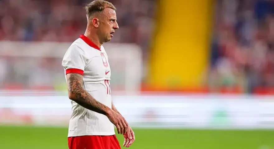 Kamil Grosicki w białej koszulce z orzełkiem i czerwonych spodenkach, gotowy do gry. Jego obecne drużyny to Polska i Pogoń Szczecin.