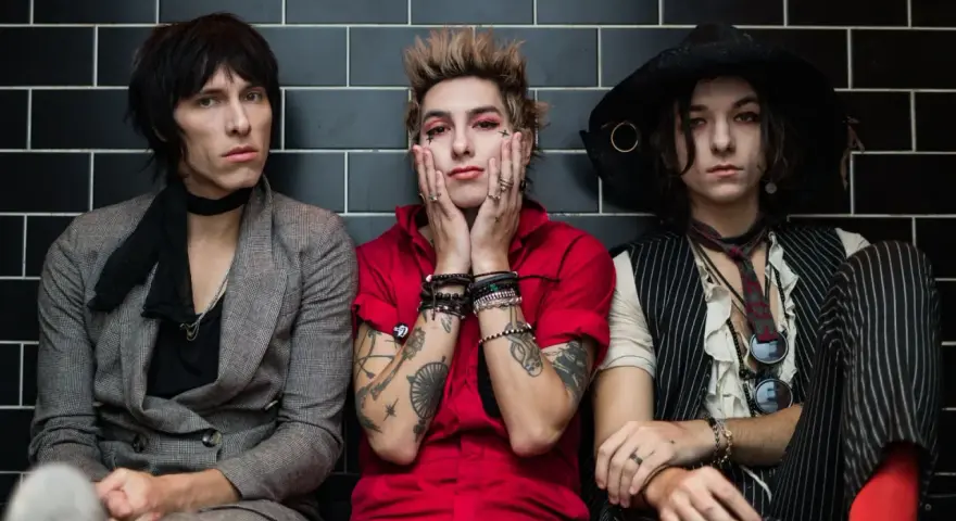 Trzech członków Palaye Royale w stylowych, rockowych strojach siedzi obok siebie na tle czarnych płytek.