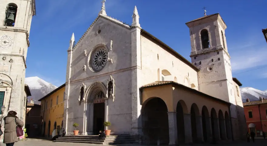 A grand church with a rose window and bell tower stands in a town square, with snow-capped mountains in the distance. This is Nursia, a historic Italian town.