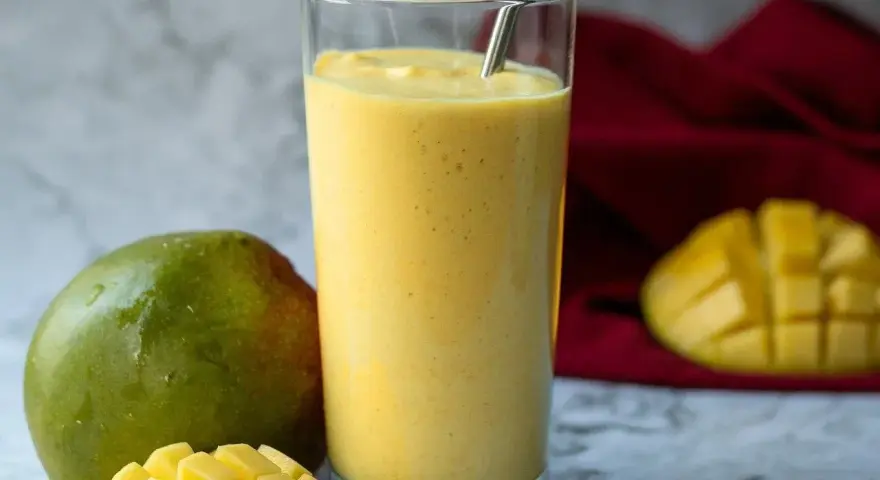 Orzeźwiający koktajl z mango w szklance, obok świeże mango i kawałki owocu. Idealny na upalne dni.