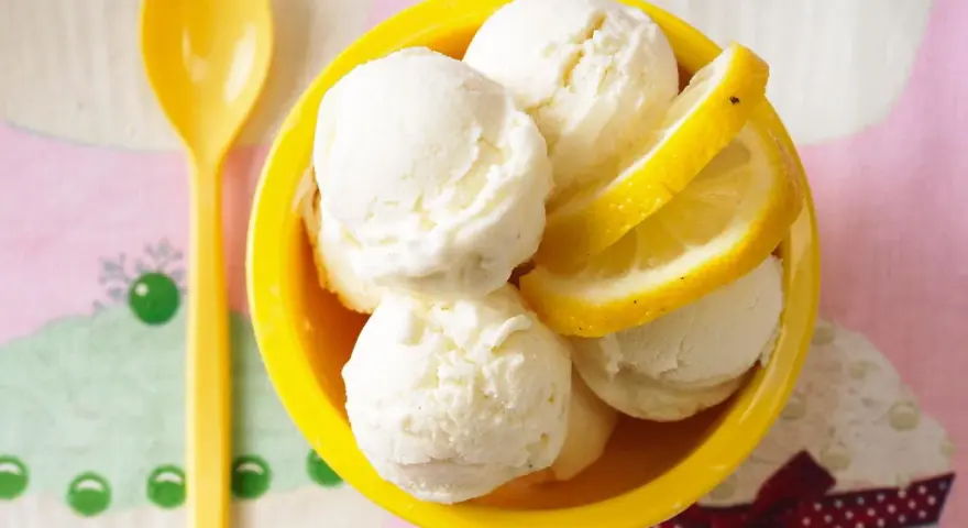 Erfrischendes Buttermilch Zitronen Eis in einer gelben Schale, garniert mit Zitronenscheiben. Ein gelber Löffel liegt daneben.