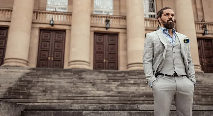 Elegancki mężczyzna w jasnej, kratkowanej marynarce i kamizelce, z chusteczką w kolorze śliwkowym w kieszeni, stoi na schodach przed budynkiem.
