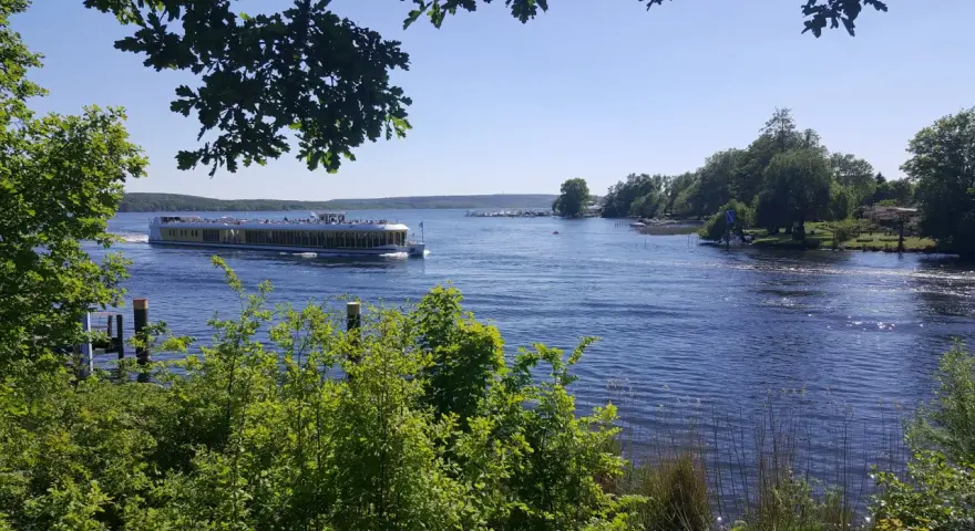 Ein Ausflugsboot fährt auf einem glitzernden See bei Potsdam. Grüne Bäume säumen das Ufer, ideal für einen entspannten Tag an den Badeseen Potsdam.