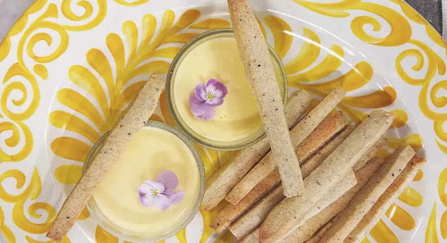 Två skålar med gul kräm, dekorerade med violer, serveras med krispiga kex på en gulmönstrad tallrik. En perfekt påsk dessert.