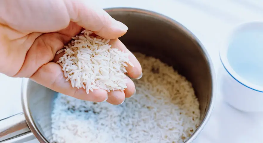 Mano sosteniendo arroz para calcular el tiempo cocción arroz perfecto. Olla con arroz y taza de agua al lado.
