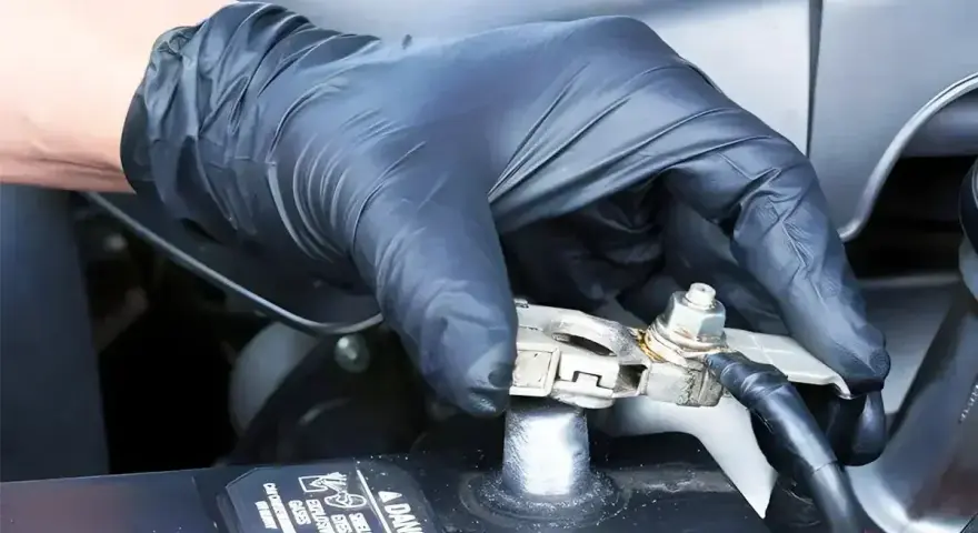 Gloved hand applying WD40 to a car battery terminal to prevent corrosion and ensure a good connection.