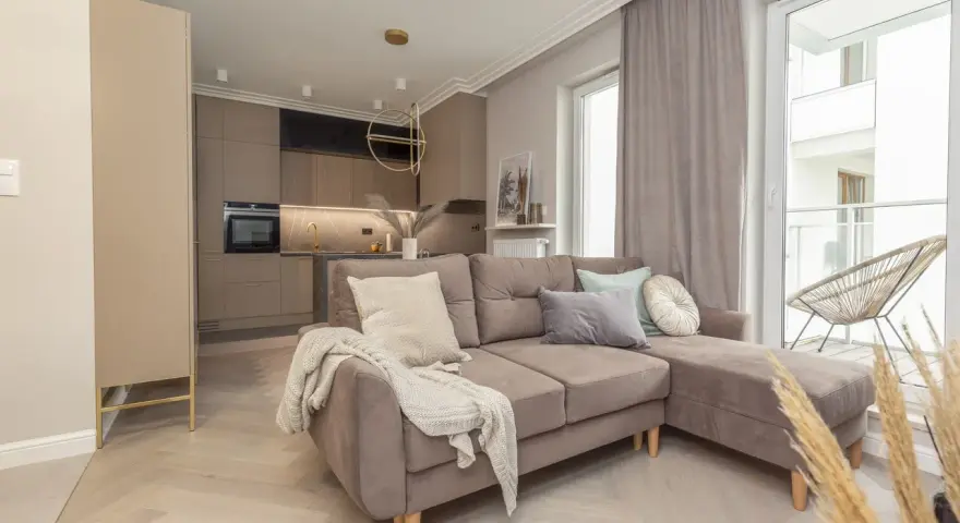 Elegancki salon z aneksem kuchennym w stylu monochromatycznym. Brązowa sofa z poduszkami, jasne drewno na podłodze.