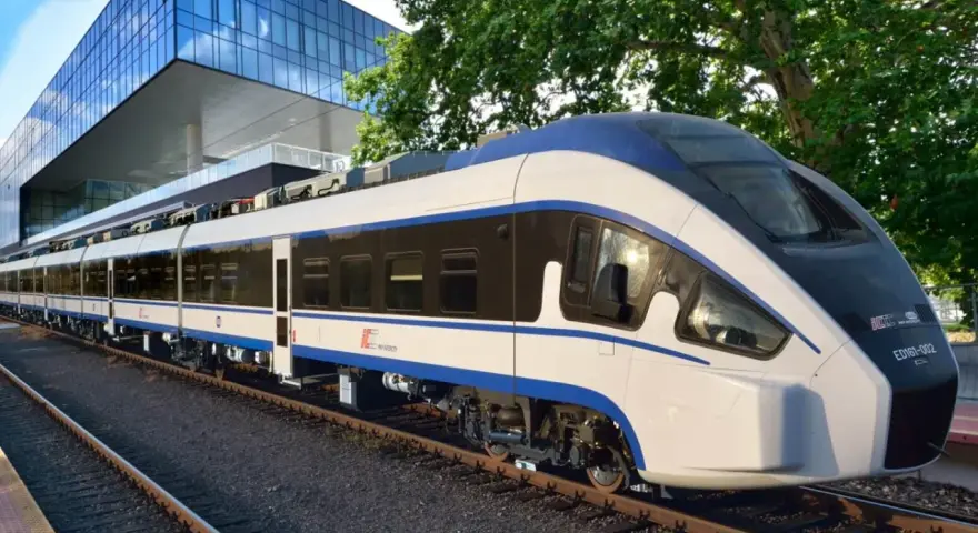 Nowoczesny, biało-niebieski pociąg Intercity ED161-002, najszybszy pociąg w Polsce, stoi na torach obok futurystycznego budynku.