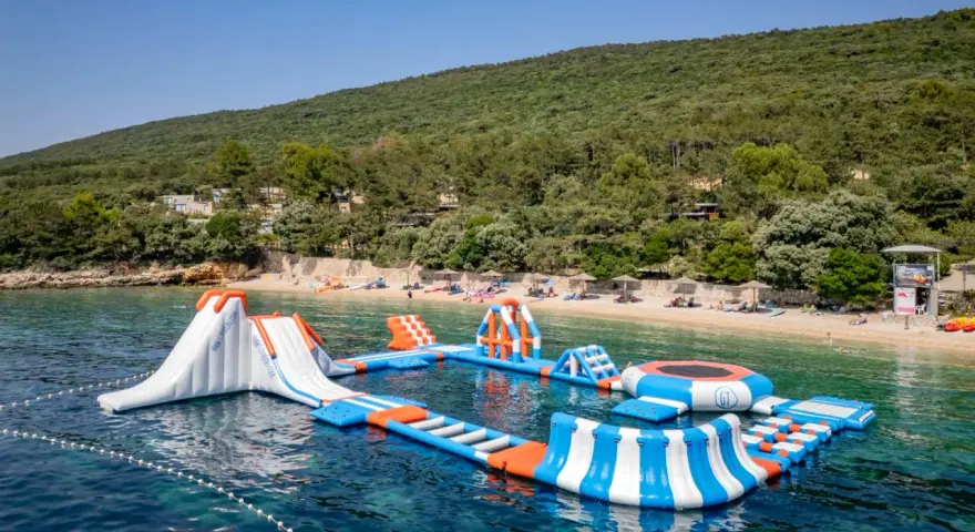 Wodne atrakcje na krystalicznie czystej wodzie, idealne na wakacje w **camping Slatina**. W tle zielone wzgórza i plaża.