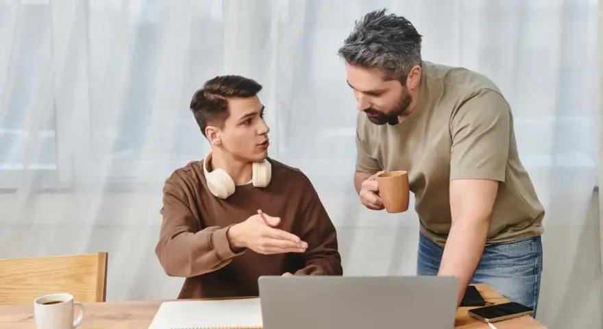 Ojciec rozmawia z synem, pokazując mu, jak zmotywować dziecko do nauki. Na stole laptop, zeszyt i kubek kawy.