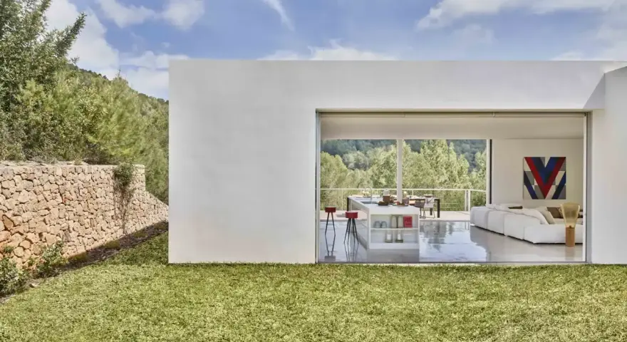 Exterior de casa ibicenca con césped verde y muro de piedra. Interior moderno con cocina y salón abiertos al paisaje.