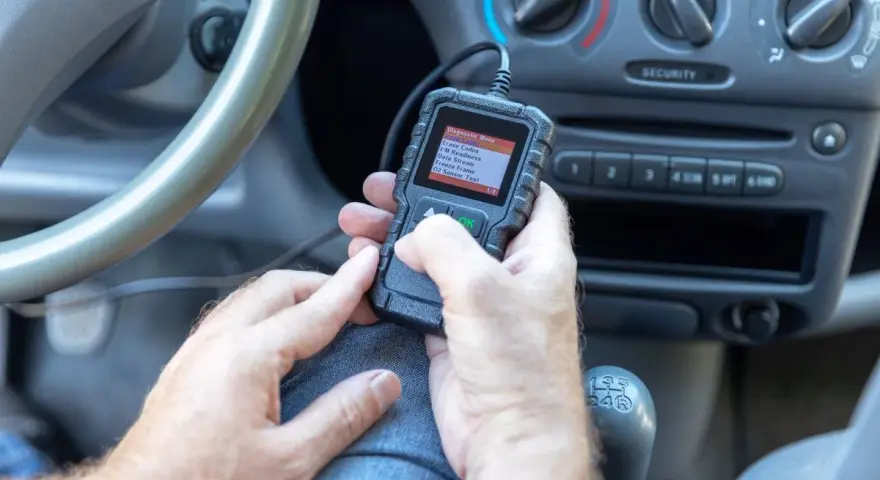 Ręce trzymające skaner diagnostyczny, który pokazuje menu z opcją "02 Sensor Test". Urządzenie może pomóc zdiagnozować problemy z czujnikiem spalania stukowego.