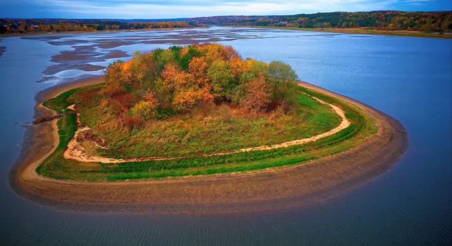 Jesienna wyspa na zalewie Brody, porośnięta kolorowymi drzewami i trawami, otoczona błękitną wodą.