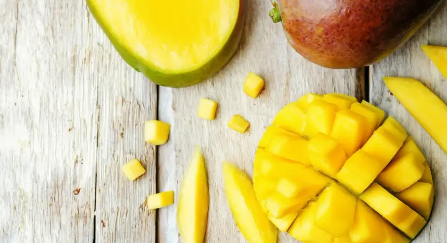 Czy pies może mango? Na drewnianym blacie leżą soczyste kawałki mango, pokrojone w kostkę i paski. Obok całe, dojrzałe owoce.
