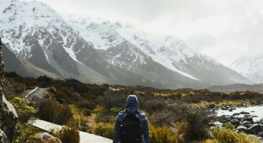 Une personne avec un sac à dos fait une randonnée sous la pluie, face à des montagnes enneigées. Le chemin en bois serpente à travers une végétation basse.