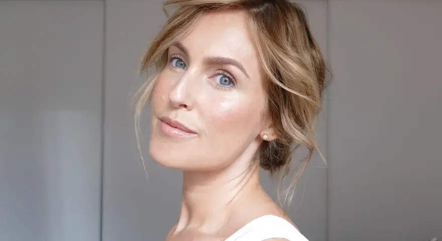 En kvinna med blont hår och blå ögon har ett naturligt glow i ansiktet. Hon bär en vit topp och en liten diamantörhänge.