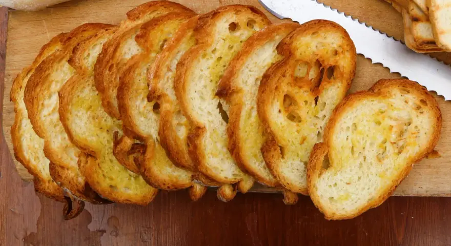 Fette dorate di pane con lievito madre, appena tostato, su un tagliere di legno. Un coltello seghettato è pronto per affettare altro pane.