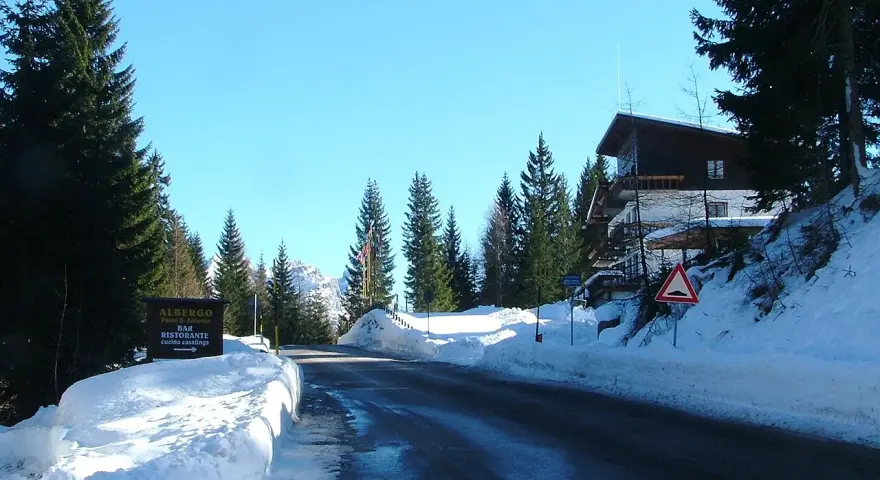 Strada innevata al passo San Antonio. Albergo e ristorante con cucina casalinga. Case di montagna e pini.