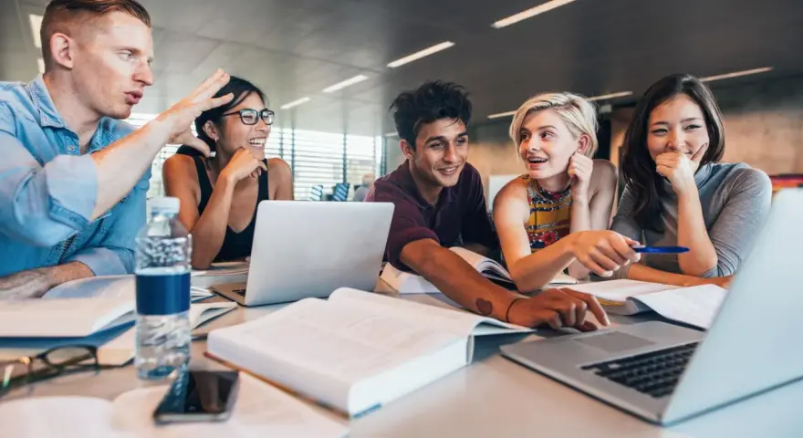 Fünf Studierende lernen gemeinsam. Sie lachen und diskutieren, während sie an Laptops arbeiten. Ein Mann gestikuliert lebhaft, was auf ein spannendes Studium im Ausland hindeutet.