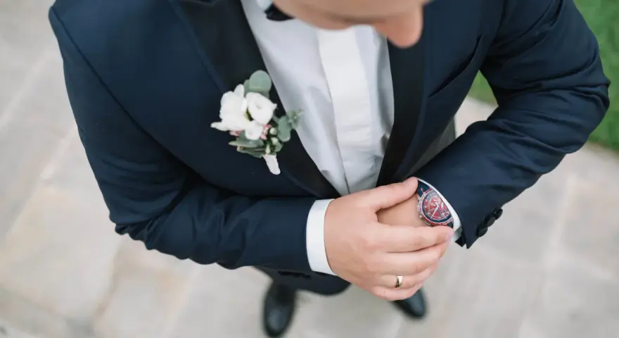 Elegancki pan młody w granatowym garniturze z butonierką co to jest, poprawia zegarek.