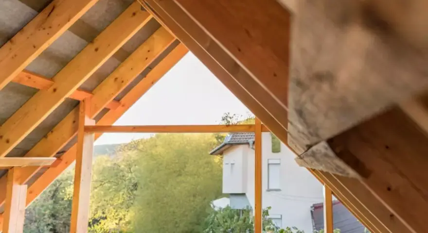 Drewniany szkielet dachu dwuspadowego w trakcie budowy. Widoczne krokwie i elementy konstrukcyjne, a w tle dom i zieleń.