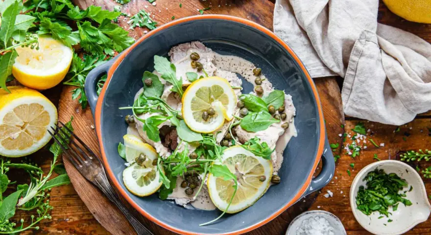 Vitello tonnato i en skål, toppad med citronskivor, kapris och ruccola. Serveras med citroner och färska örter.