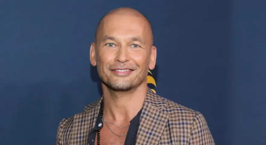 Tomasz Wygoda, łysy mężczyzna z niebieskimi oczami, uśmiecha się, ubrany w kratowaną marynarkę i naszyjnik.