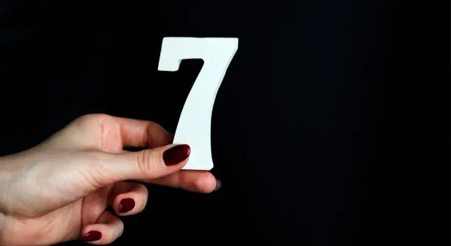 Dłoń z ciemnoczerwonym lakierem trzyma białą cyfrę 7. Może to symbolizować numerologiczną 7 mężczyzny, który jest tajemniczy i introspektywny.