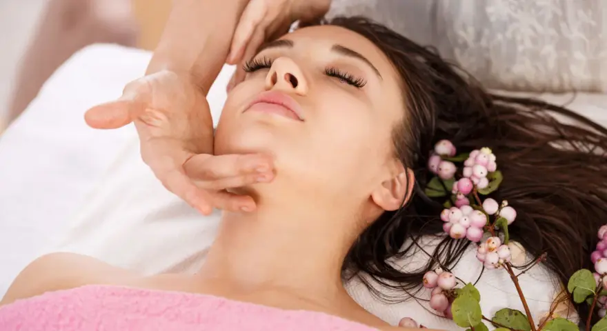 A woman receives an anti aging facial massage, her eyes closed in relaxation. Delicate pink berries adorn her dark hair.