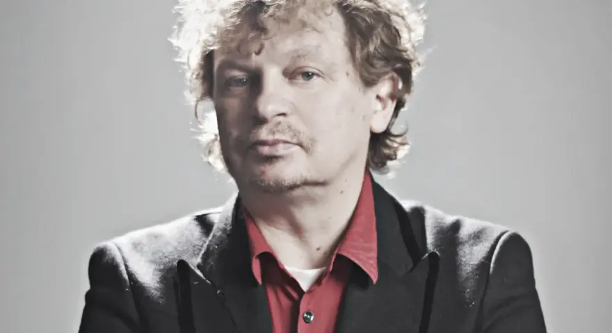 Marek Zieliński, aktor, w czarnej marynarce i czerwonej koszuli, z kręconymi włosami.