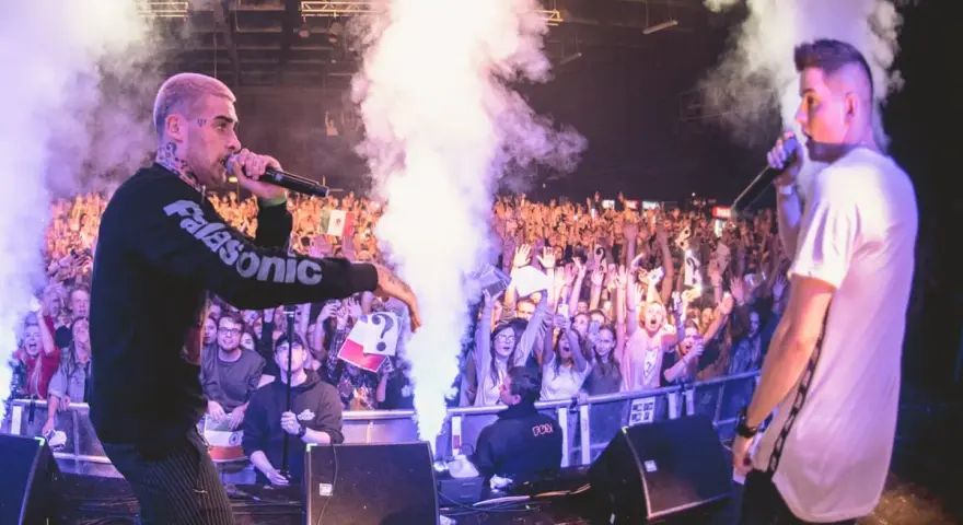 Krzysztof, w wieku swoich najlepszych lat, na scenie z mikrofonem, tłum szaleje, dym i światła tworzą atmosferę koncertu.