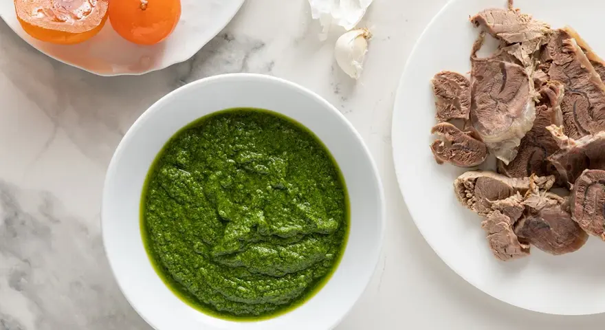Una ciotola di vibrante salsa verde accanto a fette di carne bollita e pomodorini confit.